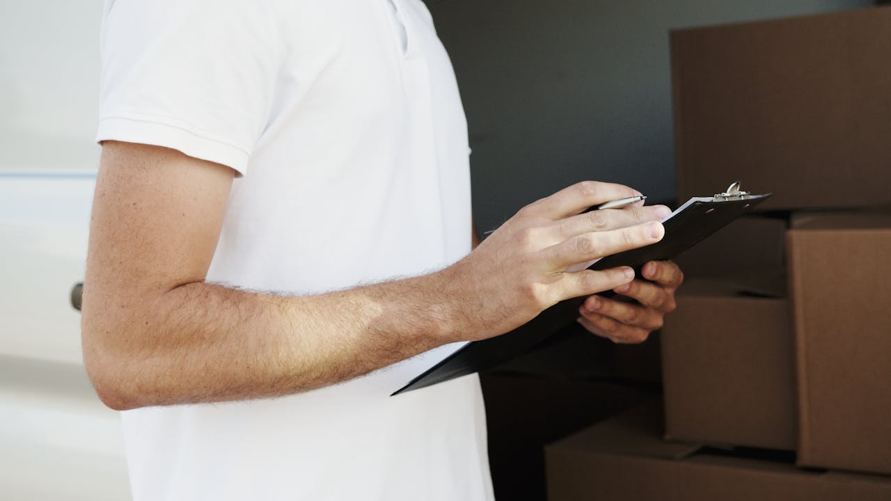A delivery person in a white shirt holding a clipboard and pen near a vehicle with boxes.